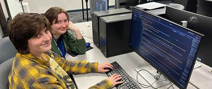 Cypher and Mickey working on this very website! Each of them have their own grey rolling chair in front of a long, white table with a set of computers, keyboards, and mice in front of each student.