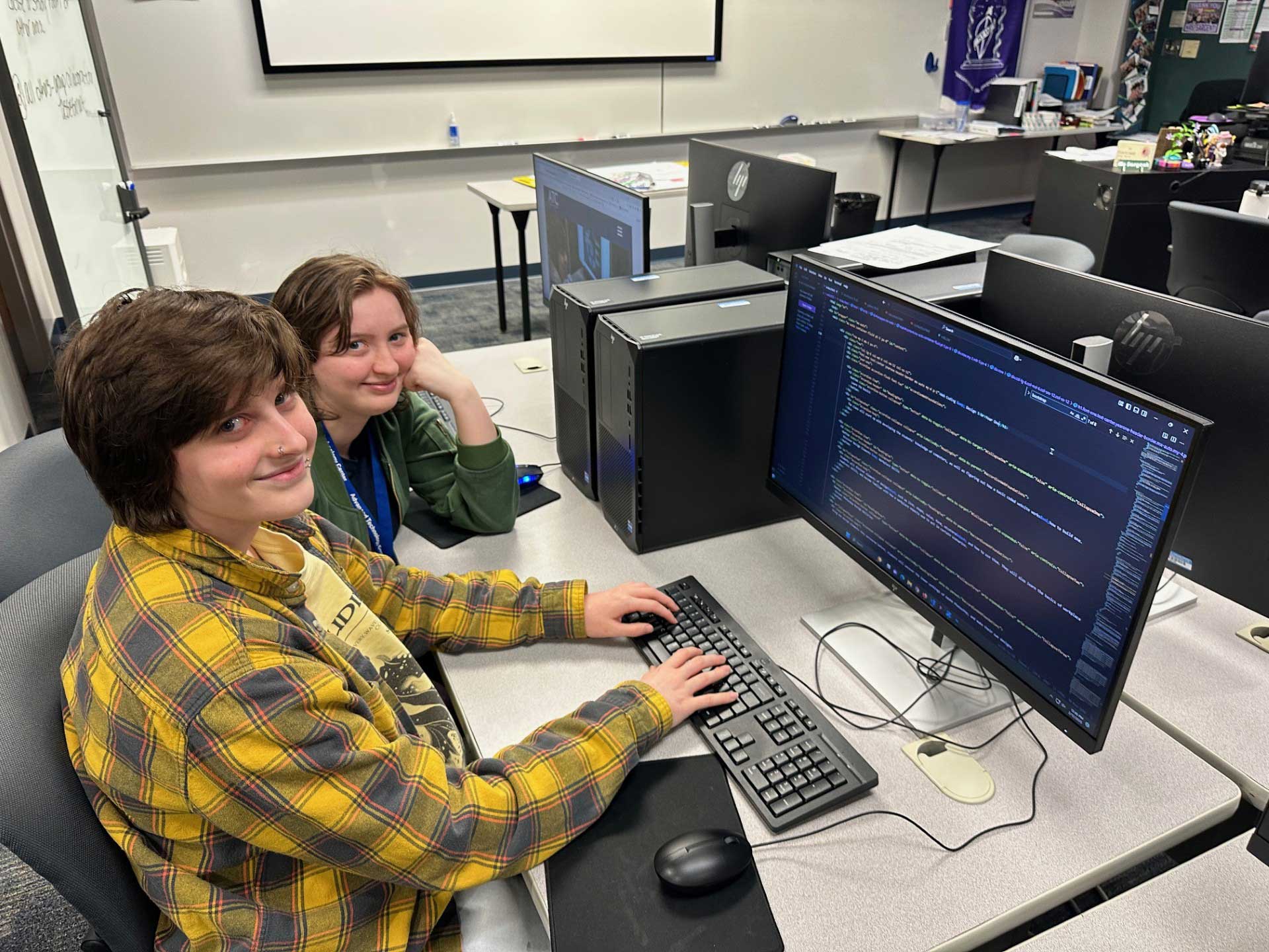 Cypher and Mickey working on this very website! Each of them have their own grey rolling chair in front of a long, white table with a set of computers, keyboards, and mice in front of each student.