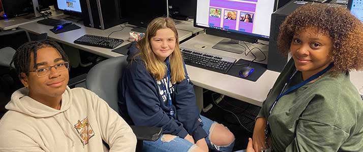 Group of students, sitting in their own grey rolling chairs at a long, white table with a set of computers, keyboards, and mice in front of each student.