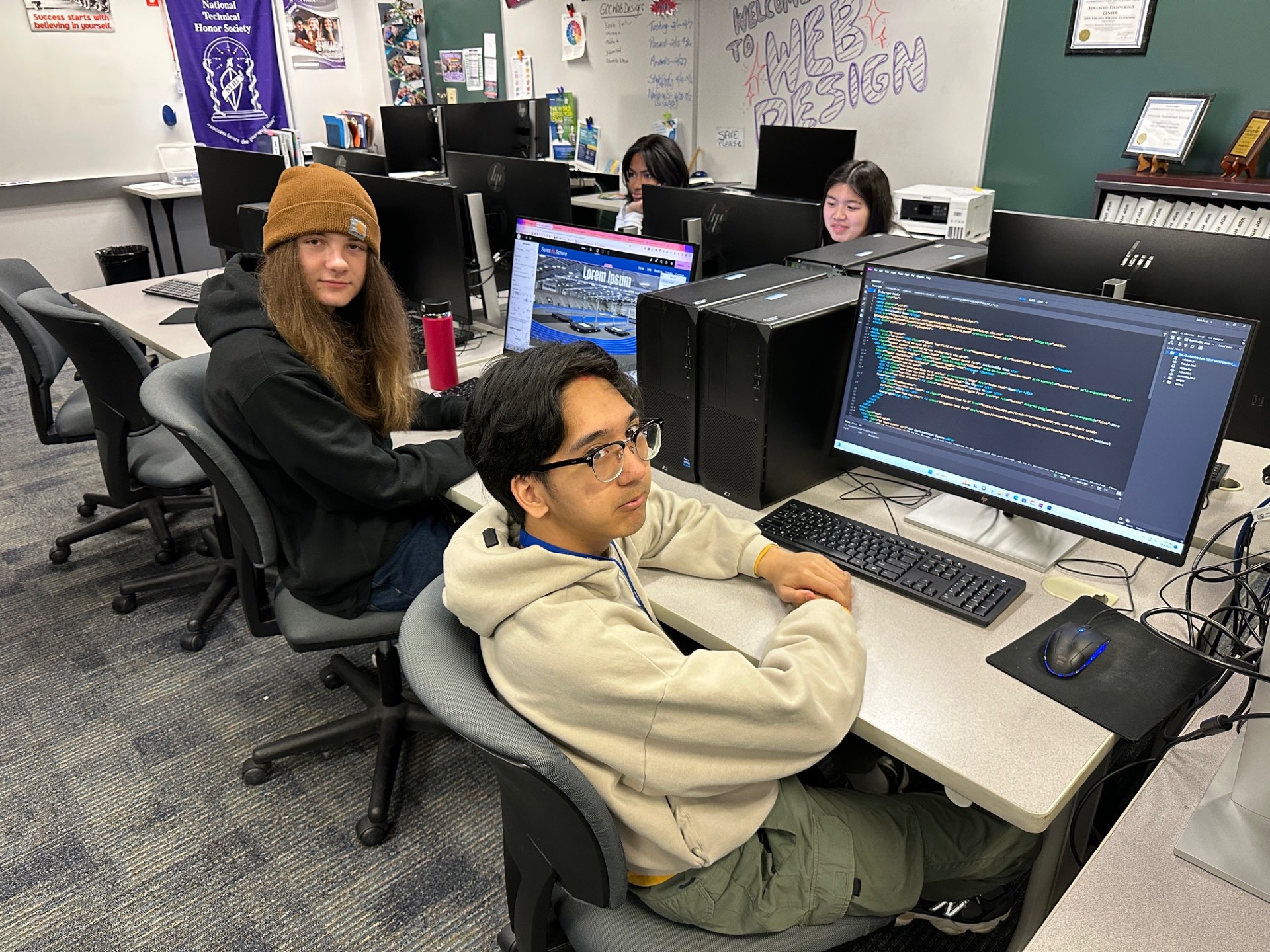 Group of students, sitting in their own grey rolling chairs at a long, white table with a set of computers, keyboards, and mice in front of each student.