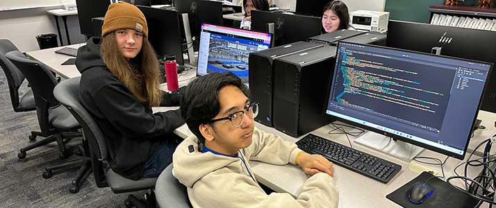 Group of students, sitting in their own grey rolling chairs at a long, white table with a set of computers, keyboards, and mice in front of each student.