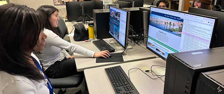 Students working on their own sites, sitting in their own grey rolling chairs at a long, white table with a set of computers, keyboards, and mice in front of each student.