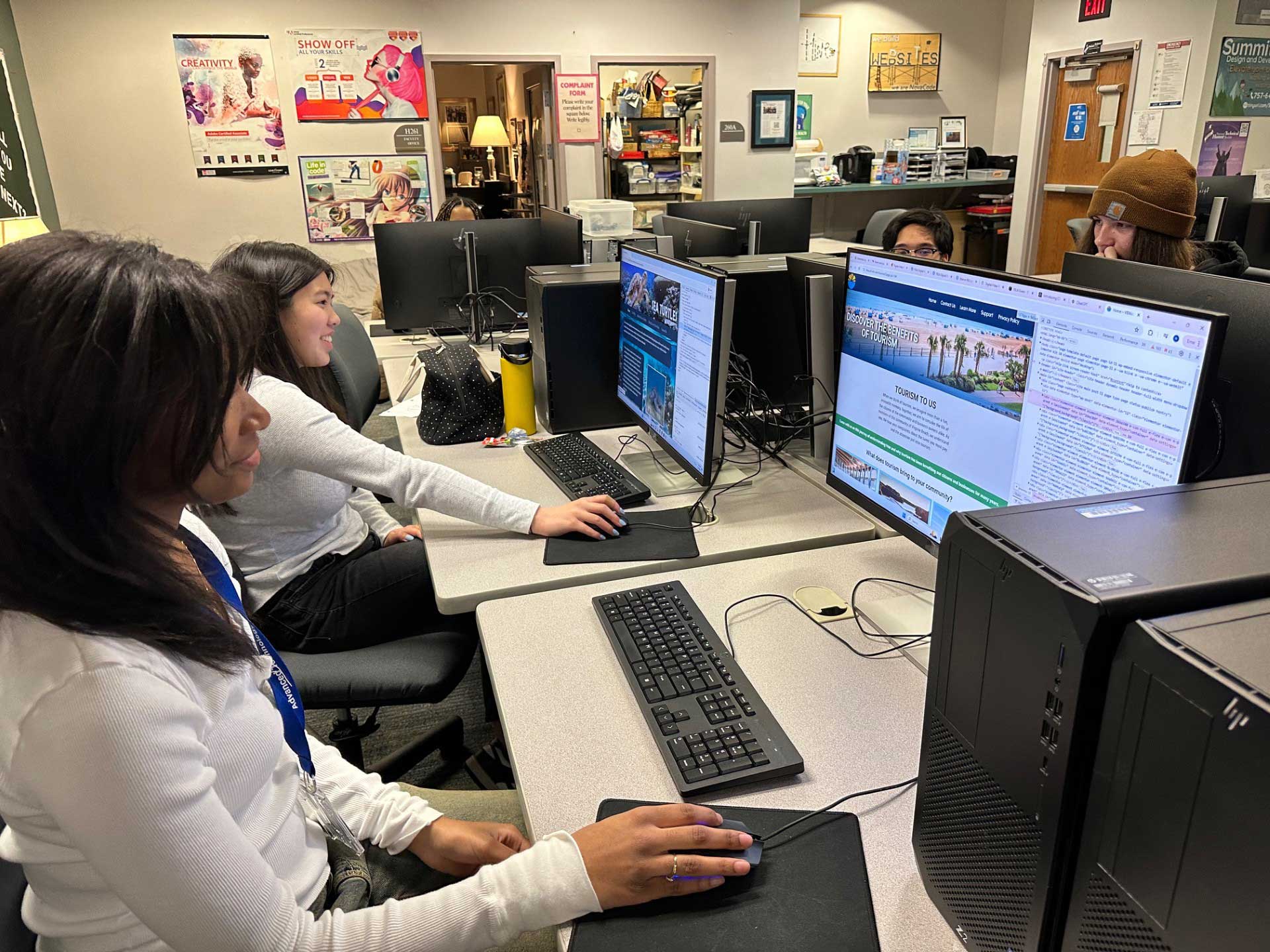 Students working on their own sites, sitting in their own grey rolling chairs at a long, white table with a set of computers, keyboards, and mice in front of each student.